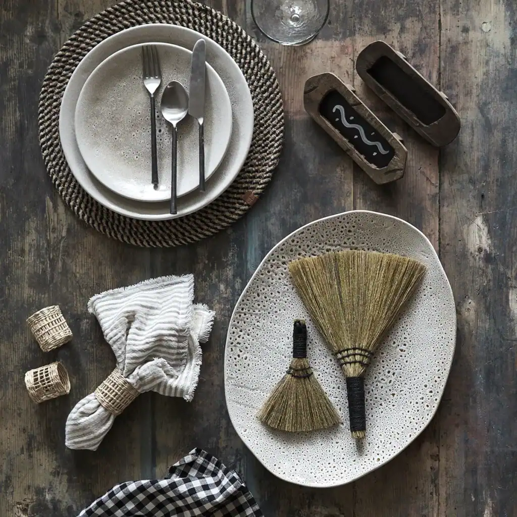 En dukning med tallrikar från serien Annabelle av Affari of Sweden. En stor och en liten beige tallrik ligger på ett rustikt träbord tillsammans med bestick, linneservetter i servettringar, och små kvastar. På bordet finns också en rund bordstablett av flätad halm, en svart- och vitrutig servett samt två trälådor.