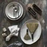 En dukning med tallrikar från serien Annabelle av Affari of Sweden. En stor och en liten beige tallrik ligger på ett rustikt träbord tillsammans med bestick, linneservetter i servettringar, och små kvastar. På bordet finns också en rund bordstablett av flätad halm, en svart- och vitrutig servett samt två trälådor.