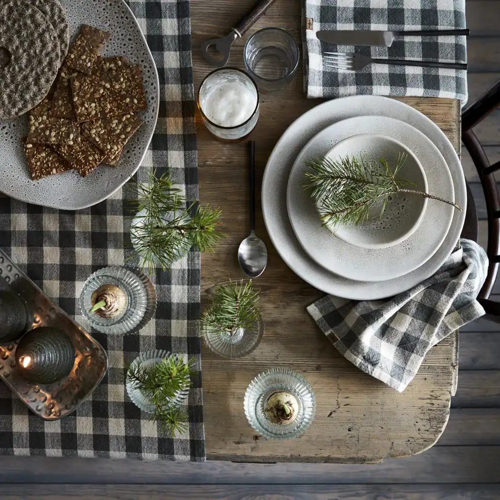 En dukning med tallrikar från serien Annabelle av Affari of Sweden. Tallrikarna är staplade och dekorerade med en grankvist. På bordet finns även en ölglas med skum, en sked, krispbröd på en tallrik, och glasbehållare med små grankvistar. Bordet är täckt med en svart- och vitrutig duk och servett.