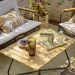 Bloomingville Hampton utebord i akaciaträ, elegant presenterat på en solig terrass med sittplatser, drycker och frukter, perfekt för avkopplande stunder utomhus.