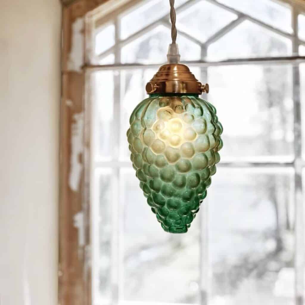 Närbild på Taklampa DRUVA från Strömshaga, tänd, med en grön glasskärm och mässingsfäste framför ett fönster.