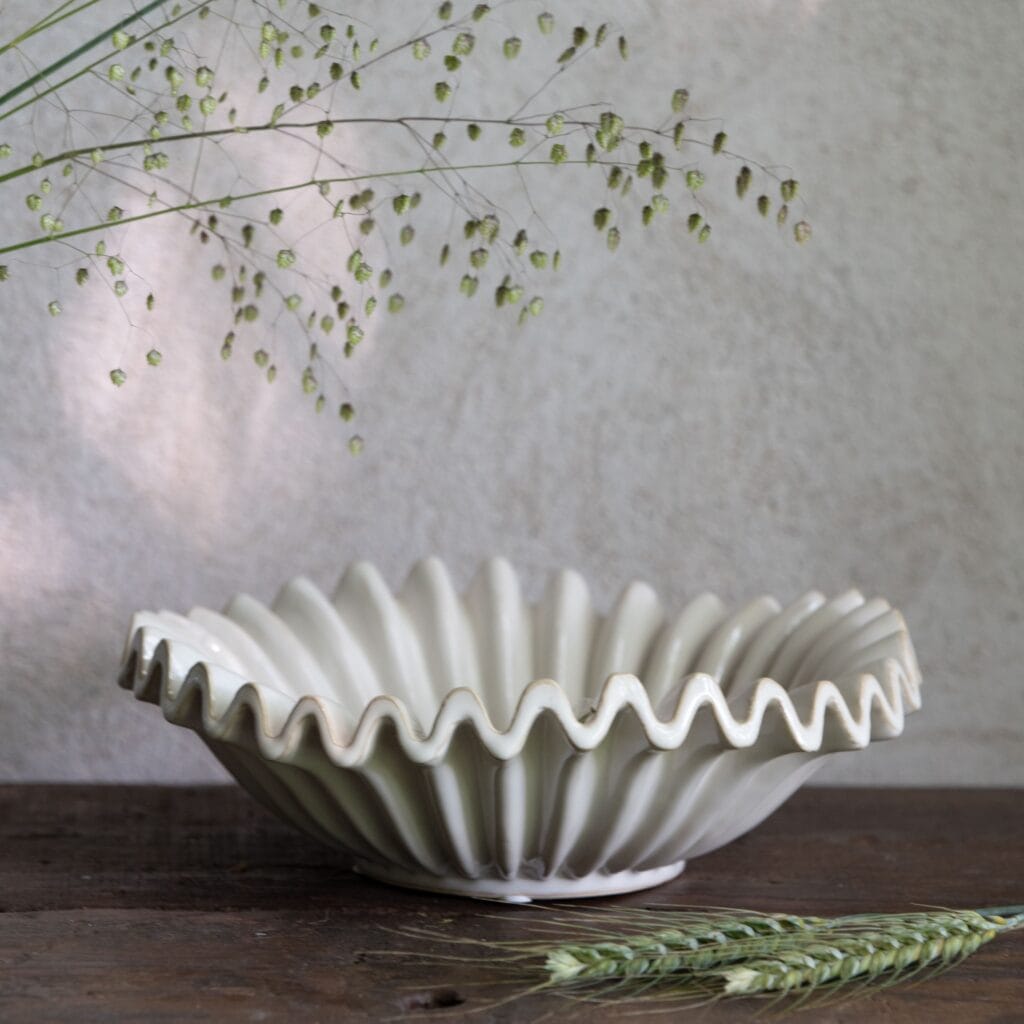 Vitmelerat stengodsfat HANNA från Wikholm Form, placerat på ett rustikt träbord med dekorativa gröna kvistar.