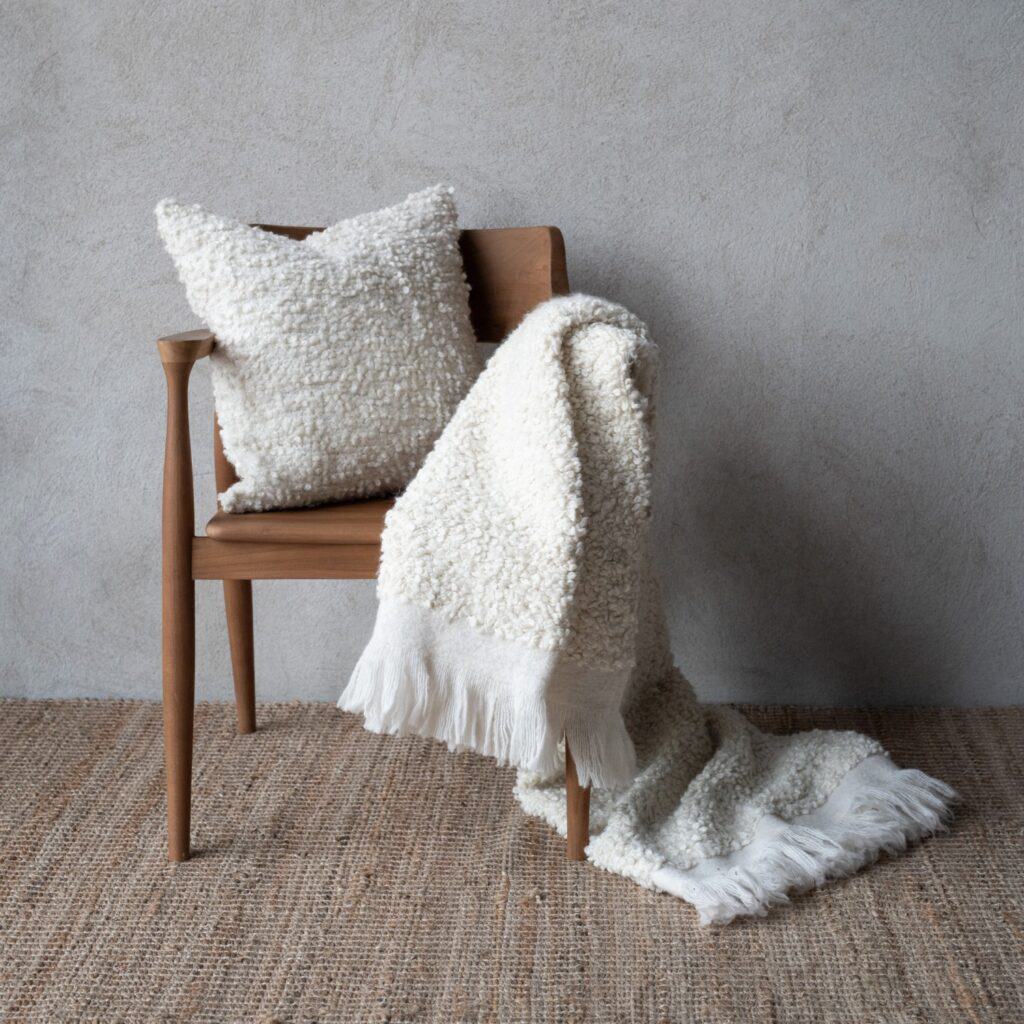 Offwhite pläd NALIA från Wikholm Form, draperad över en trästol tillsammans med ett matchande kuddfodral, mot en neutral vägg.