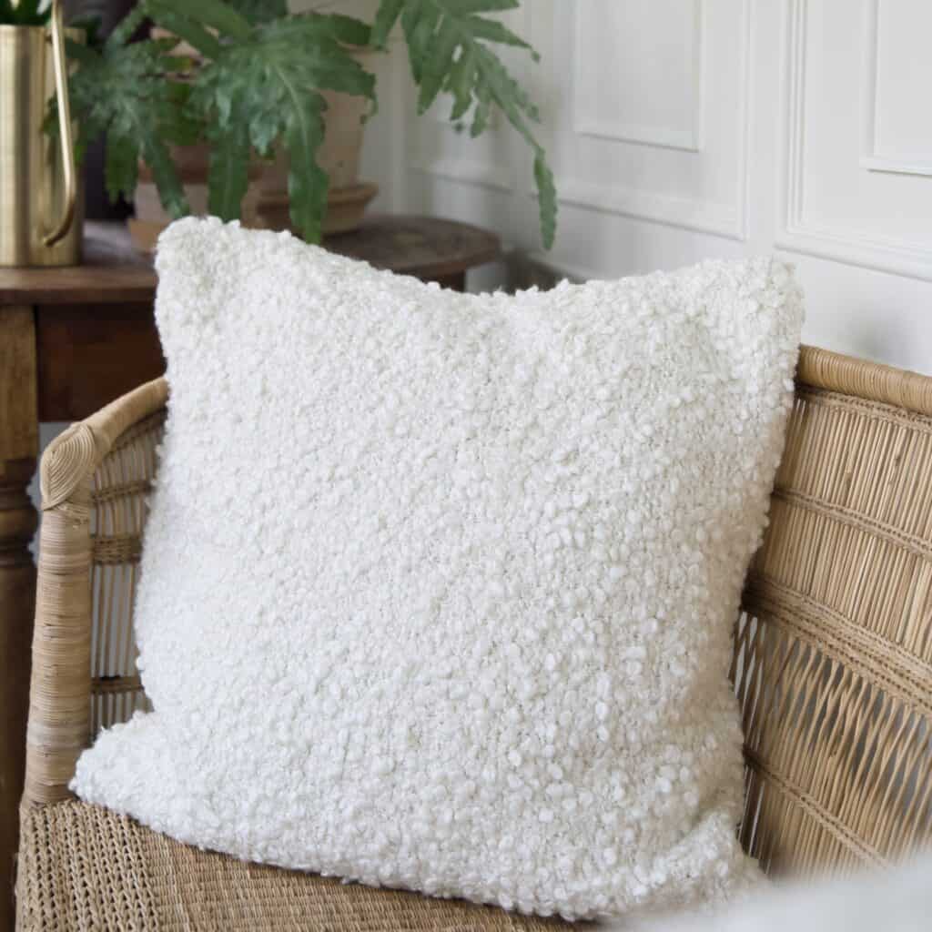 Vackert kuddfodral i vitt, NALIA, i en rottingsoffa och gröna växter i bakgrunden.