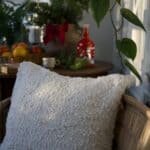 Kuddfodral NALIA i offwhite boucle på rottingstol i vinterljus, med juligt stilleben i bakgrunden.