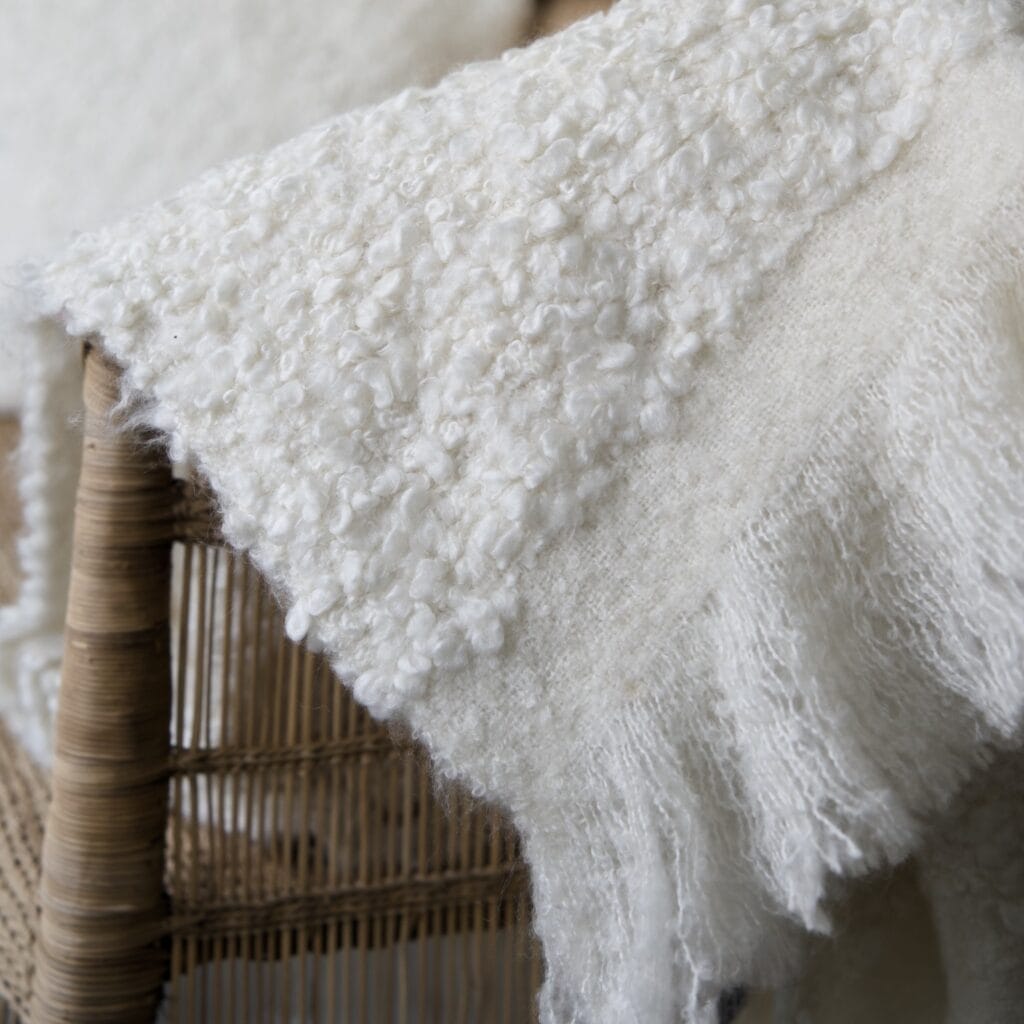 Närbild på pläden NALIA från Wikholm Form i mjuk, vit bouclé. Pläden hänger över en rottingstol och visar tydligt den texturerade ytan och de mjuka fransarna längs kanten.