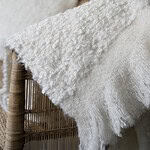 Närbild på pläden NALIA från Wikholm Form i mjuk, vit bouclé. Pläden hänger över en rottingstol och visar tydligt den texturerade ytan och de mjuka fransarna längs kanten.