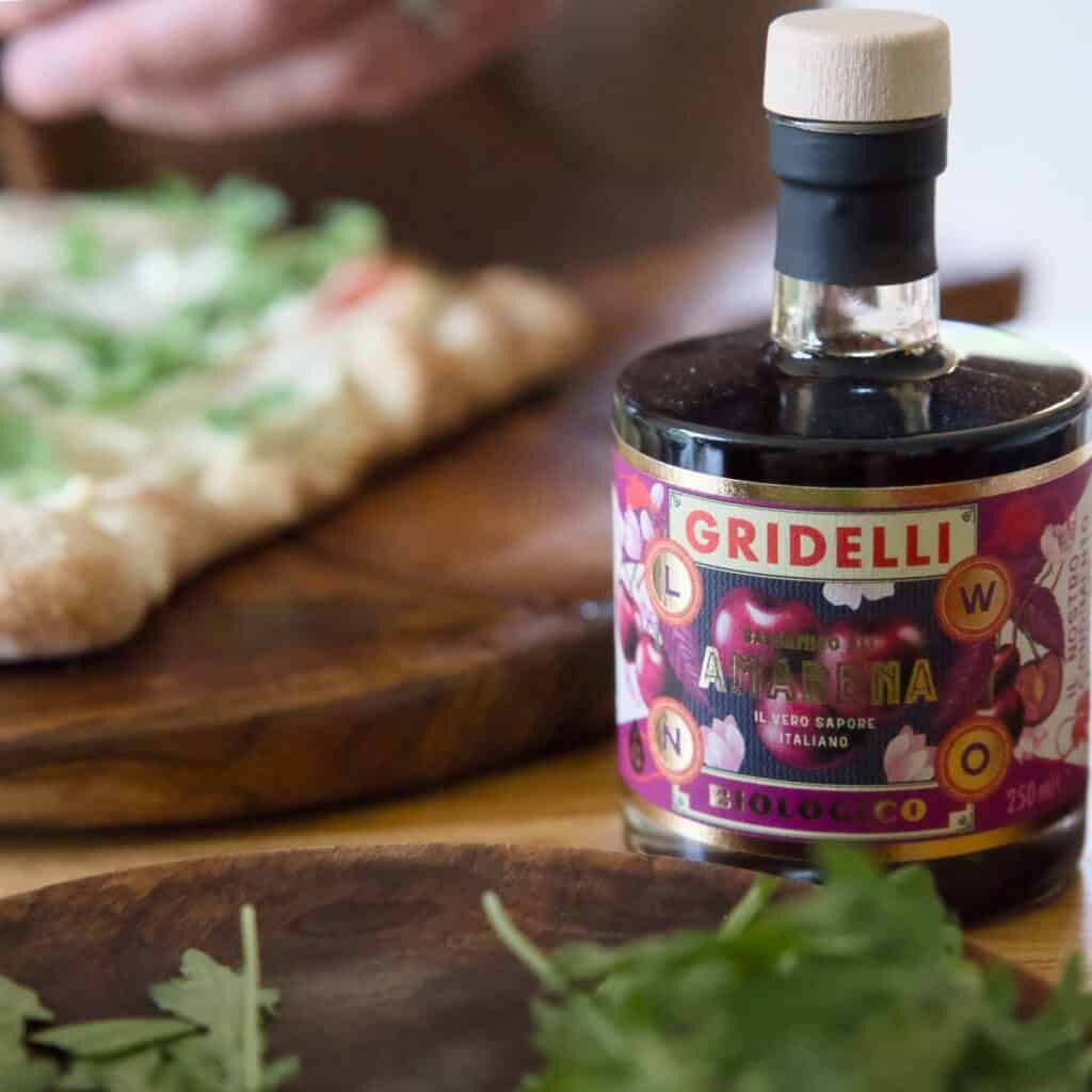 Närbild av en flaska Gridelli Balsamico Amarena, placerad på ett bord bredvid en träskärbräda med färska örter och en pizza med gröna blad. Balsamvinägern har en etikett med körsbär och andra dekorativa element.