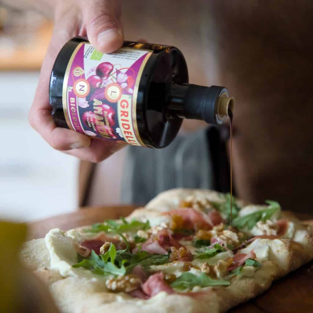 En person häller Gridelli Balsamico Amarena över en pizza toppad med ruccola, prosciutto och valnötter.