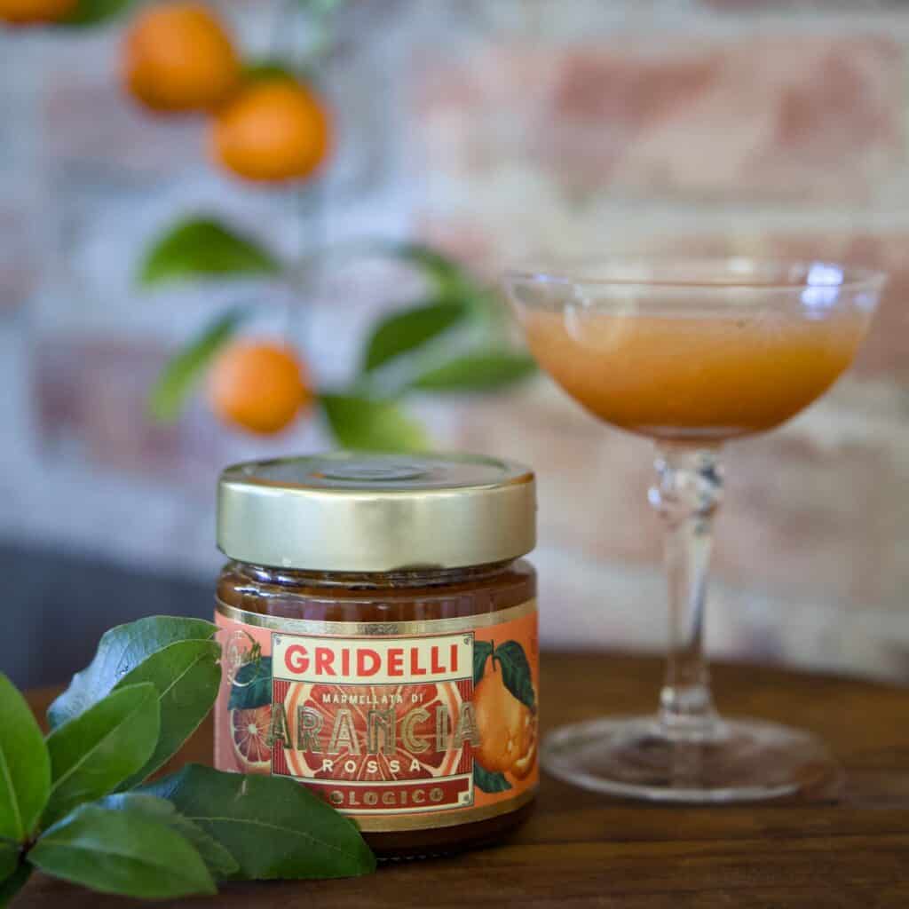 En burk ekologisk blodapelsinmarmelad från Gridelli står på ett rustikt träbord, omgiven av gröna citrusblad. I bakgrunden syns ett cocktailglas fyllt med citrusjuice och en apelsinväxt mot en tegelvägg.