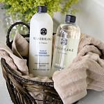 Washologi tvättmedel och mjukmedel i en korg med beiga handdukar och gröna växter i bakgrunden.