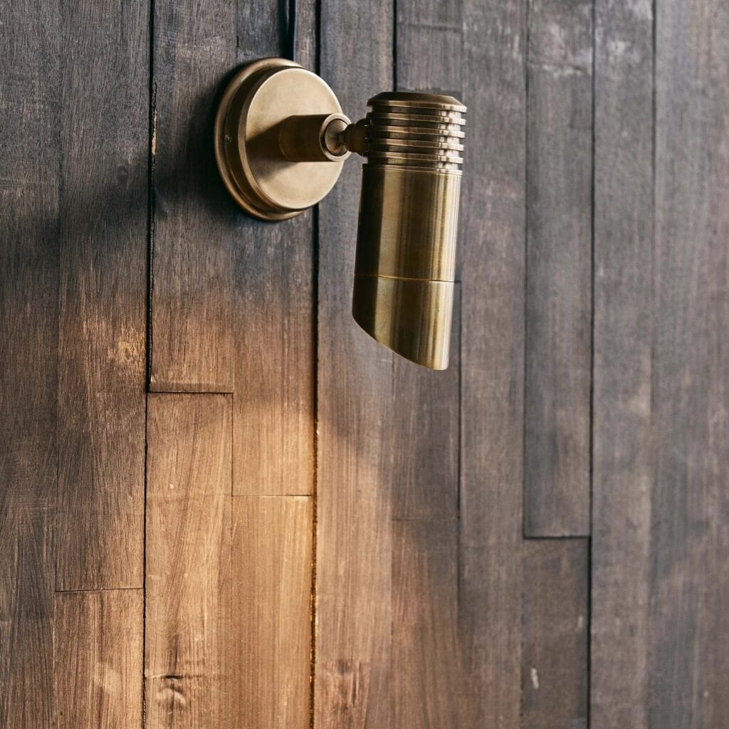 Bragi vägglampa från Nordal i mässing, monterad på en mörk trävägg. Elegant utebelysning med justerbar spot som åldras vackert med patina över tid.