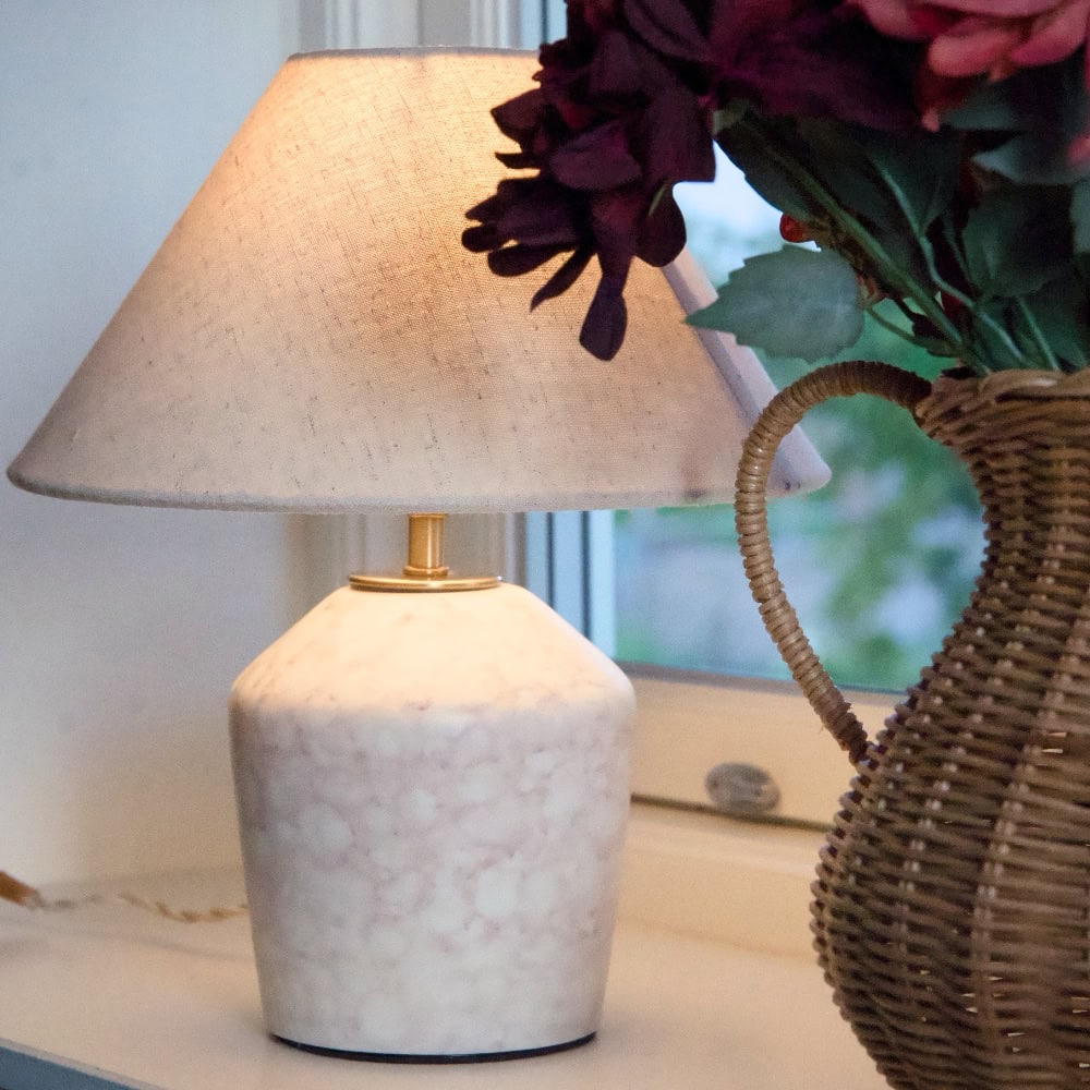 Bordslampa BODIL med marmorerad fot i beige och tänd lampskärm på en fönsterbräda bredvid en rottingvas med blommor.