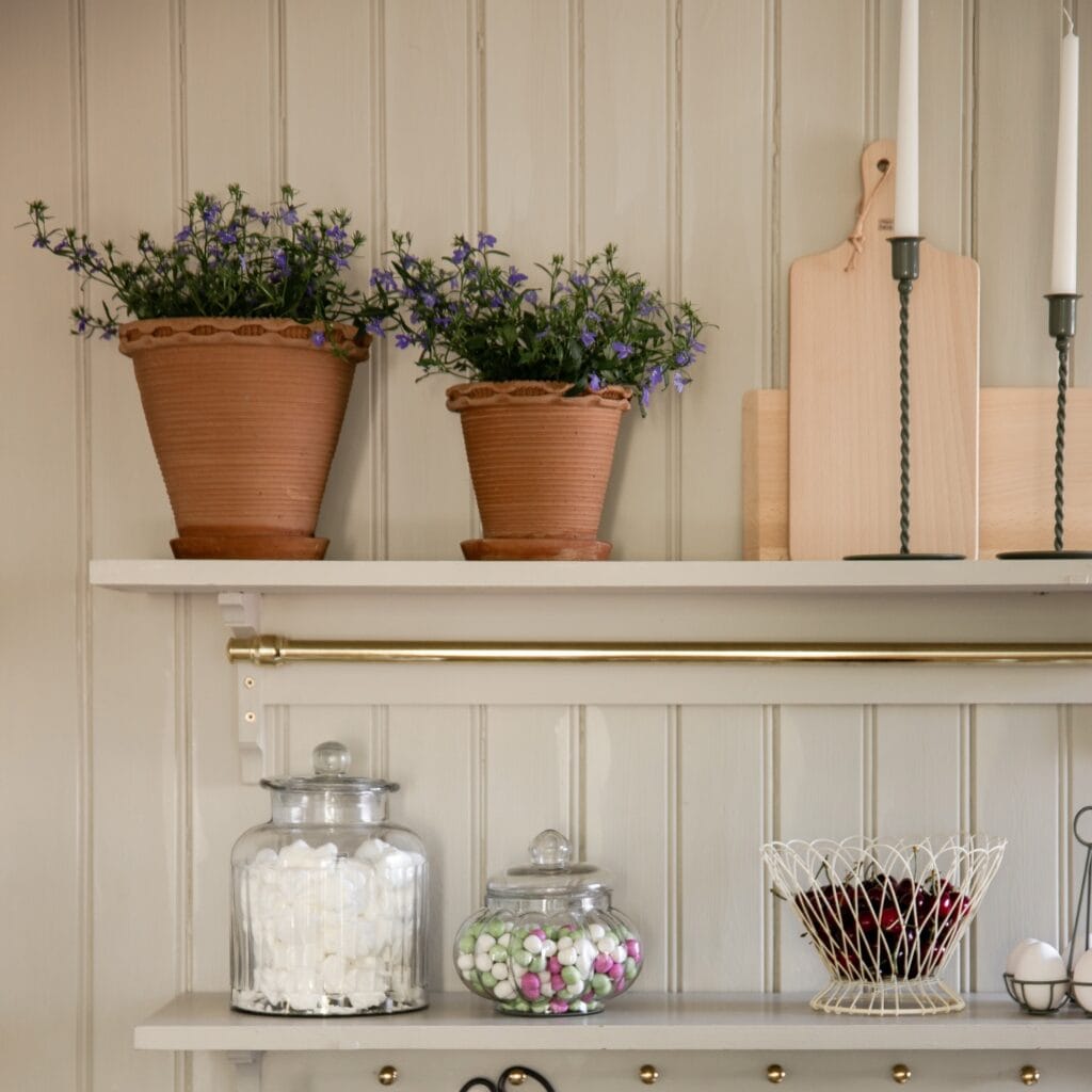 Två Alvaro-krukor med blommor, placerade på en vit hylla i ett rustikt kök med glasburkar.