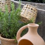 Närbild på ljusslinga i bambu från Madam Stoltz, upphängd framför en rosmarinplanta i terrakottakruka, med en handgjord ljuslykta i lerkrus i förgrunden.