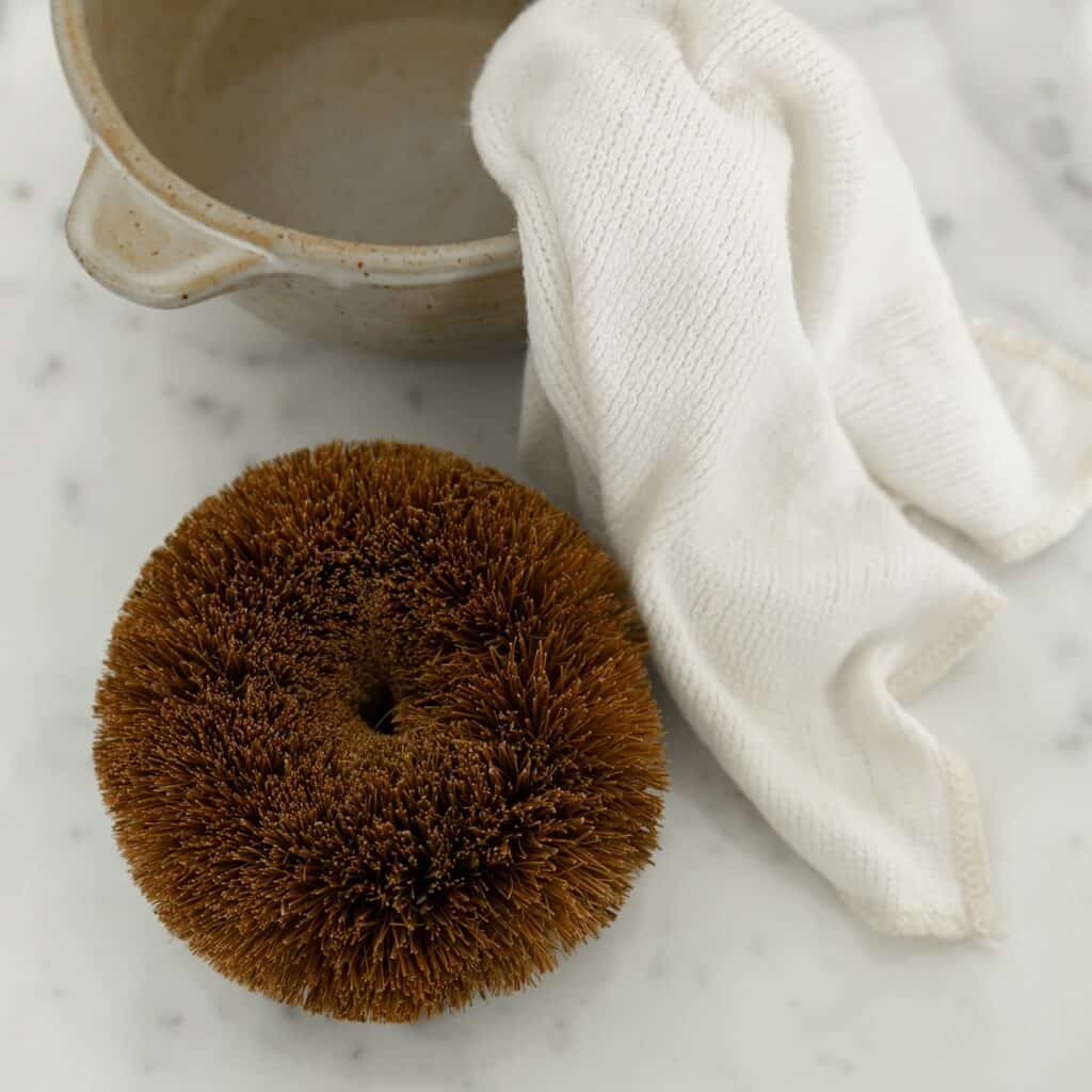 Skrubboll av kokosnötsfiber placerad bredvid en vit disktrasa och en skål på ett marmorbord.