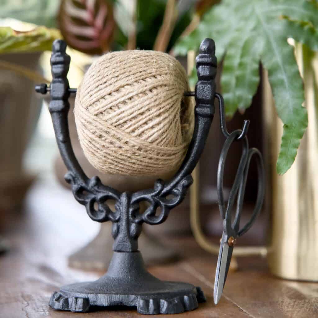 Närbild på en dekorativ snörhållare i svart gjutjärn med jutesnöre och sax, placerad på ett bord med växter i bakgrunden.