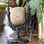 Närbild på en dekorativ snörhållare i svart gjutjärn med jutesnöre och sax, placerad på ett bord med växter i bakgrunden.