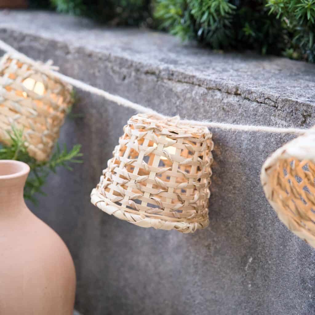 Tänd ljusslinga i handflätad bambu från Madam Stoltz, upphängd mot en stenvägg i kvällsljus, med varm glöd inuti de små skärmarna.