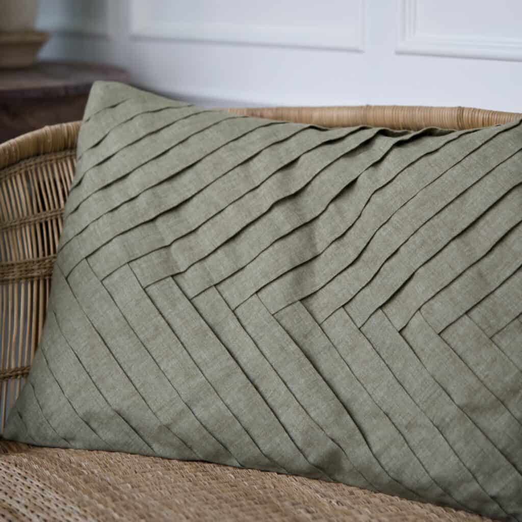 Kuddfodral FERNANDO i olivgrönt bomullstyg med ett elegant diagonalt mönster, placerat på en rottingmöbel.
