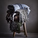 Stapel av FRASSE-filtar i olika färgnyanser på en rustik träpall, mot en mörk bakgrund som framhäver de naturliga färgerna.