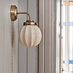 Vägglampan Marit från Strömshaga på en grå trävägg i ett rum med färgade fönster. Lampans opalglas reflekterar ljuset i rummet.