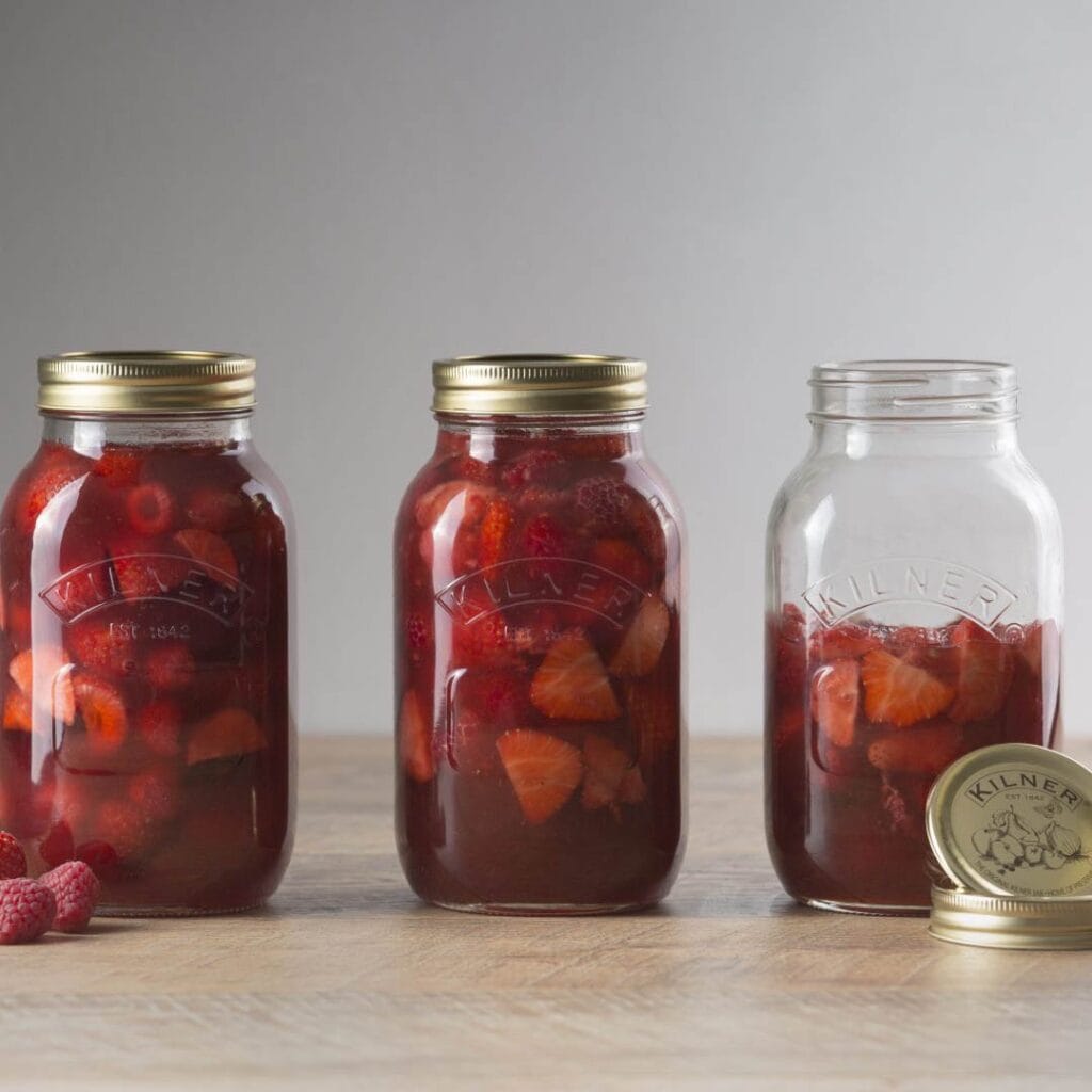 Tre Kilner konserveringsburkar fyllda med inlagda bär, två med guldfärgade lock och en öppen utan lock.
