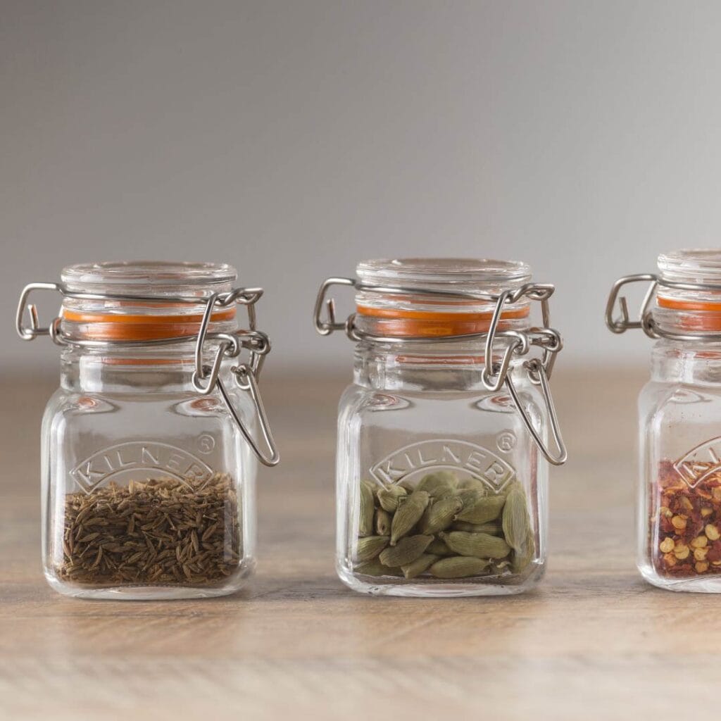 Tre Kilner bygelburkar på rad, fyllda med kryddor som kummin, kardemumma och chili, för elegant och praktisk köksförvaring.