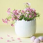 Äggformad kruka Mona i vitmelerad design, fylld med rosa blommor och stylad mot en gul bakgrund, perfekt för påskdekoration och vårens inredning.