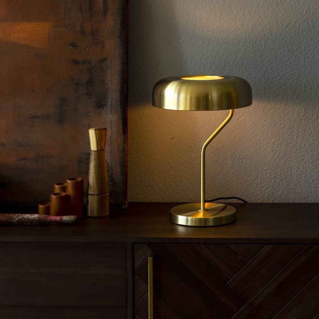Bordslampa ECLIPSE i mässing från Dutchbone, placerad på en mörk träbyrå med mjukt ljus som skapar en stämningsfull miljö.