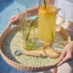 Hand som håller en rund mosaikbricka med solrosmotiv, fylld med glas, en karaff med lemonad och underlägg, placerad vid en pool.