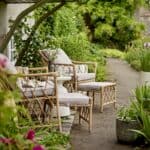 Två Belluce fåtöljer med fotpallar från Bloomingville placerade i en grönskande trädgård med blommor och stigar, stylade med beige dynor och ett litet sidobord.