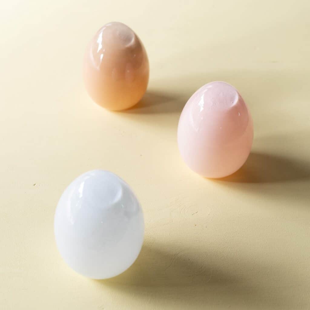 Tre glasägg i vitt, rosa och sand från Wikholm Form, placerade på en ljusgul bakgrund för en stilren och vårig känsla.