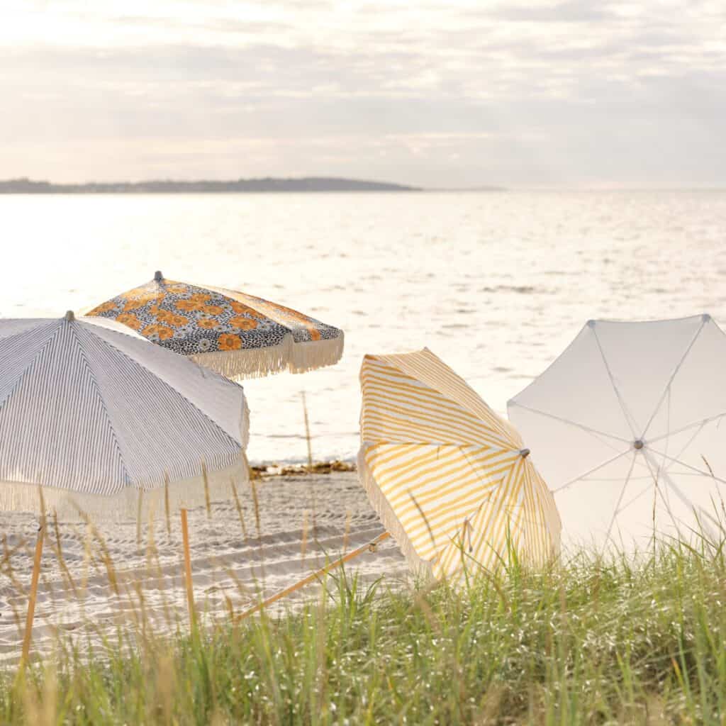 Flera parasoller från Brafab i olika färger och mönster, inklusive grårandigt, gult och blommigt, uppsatta på en solig sandstrand vid havet.