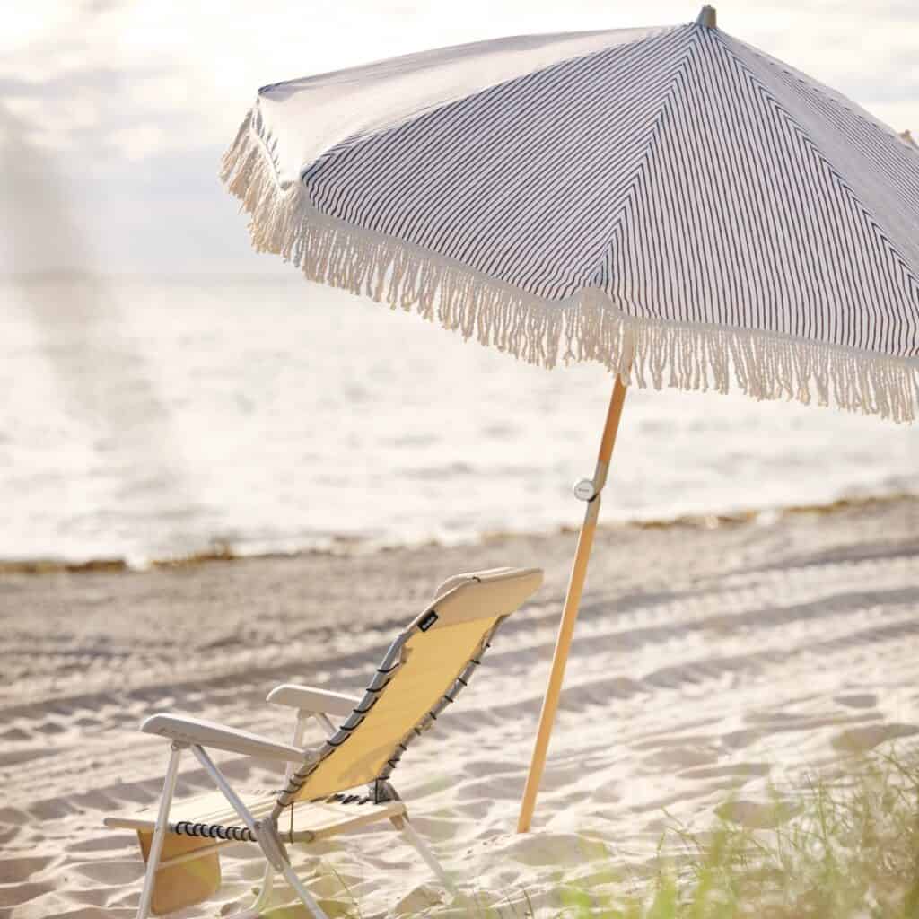 Gatsby parasoll från Brafab i grårandigt tyg med fransar, uppsatt på en sandstrand bredvid en vilstol, med havet i bakgrunden.