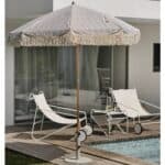Närbild på Gatsby parasoll från Brafab med grårandigt tyg och fransar, monterat i en parasollfot vid en pool och vita solstolar.