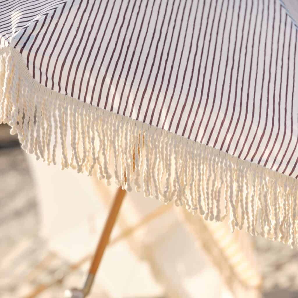 Närbild på grårandigt tyg och dekorativa fransar från Gatsby parasoll från Brafab, upplyst av solen på en strand.