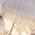 Närbild på grårandigt tyg och dekorativa fransar från Gatsby parasoll från Brafab, upplyst av solen på en strand.