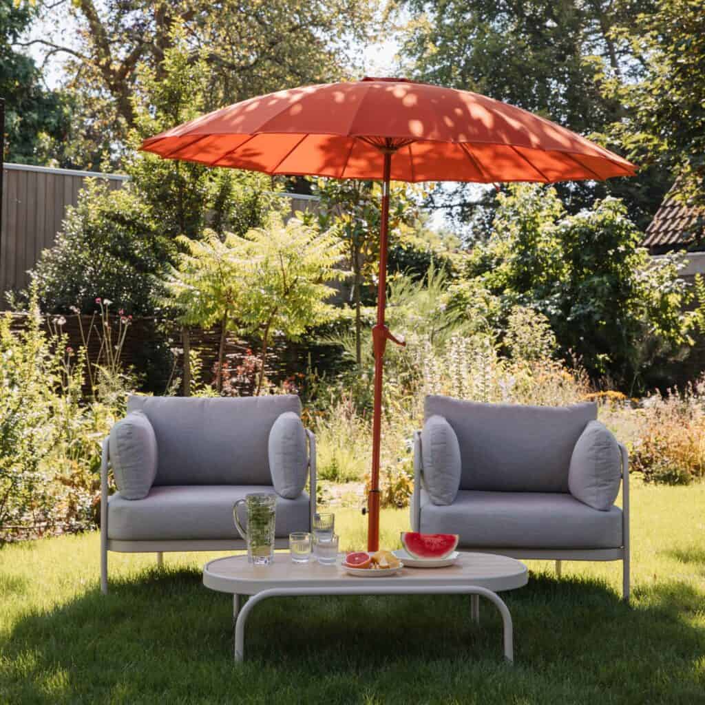 Marsalarött parasoll från Zuiver placerat i en grönskande trädgård, omgiven av två gråa loungestolar och ett litet bord med frukt och dryck.