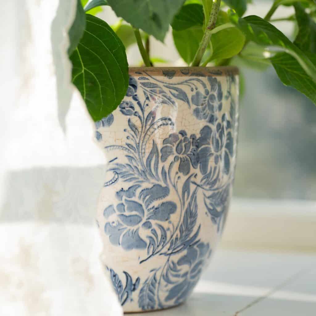 Närbild av Mirabelle-kruka i blåvitt stengods med blommönster från Wikholm Form, stående i ett soligt fönster med en grön växt.