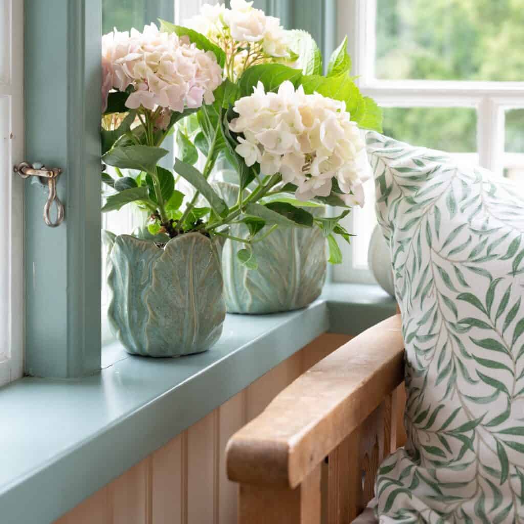 Kuddfodral Fay med grönt bladmönster placerat på en träbänk vid ett fönster med hortensior i gröna keramikkrukor.