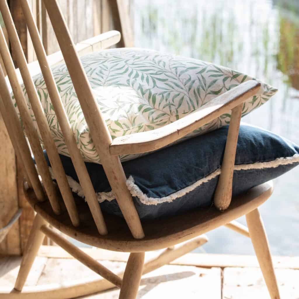 Kuddfodral Fay med grönt bladmönster placerat på en blå kudde i en rustik trästol vid vattnet.