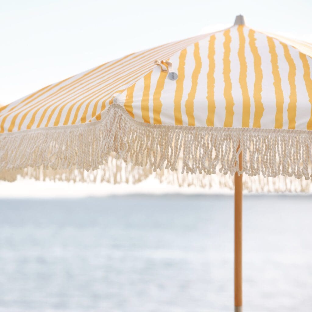 Närbild på Gatsby parasoll i gul- och vitrandigt mönster med dekorativa fransar, uppsatt vid en solig strand med havet i bakgrunden.