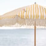 Närbild på Gatsby parasoll i gul- och vitrandigt mönster med dekorativa fransar, uppsatt vid en solig strand med havet i bakgrunden.