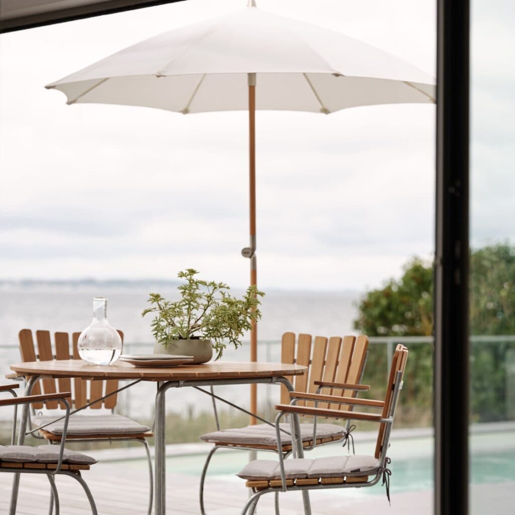 Gatsby parasoll i naturbeige från Brafab på en terrass med utsikt över havet, skapar en skuggig och avslappnande matplats med träbord och stolar.