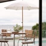 Gatsby parasoll i naturbeige från Brafab på en terrass med utsikt över havet, skapar en skuggig och avslappnande matplats med träbord och stolar.