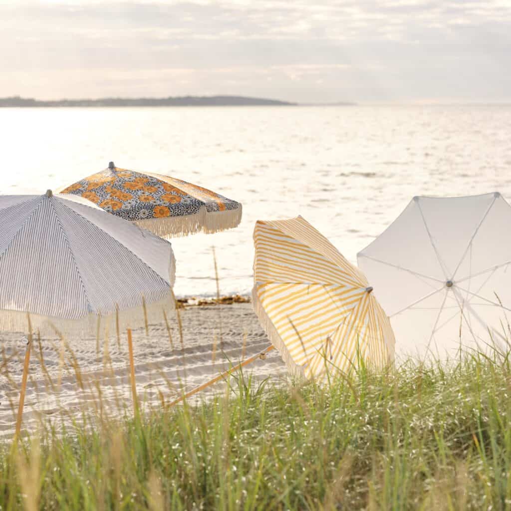 Fyra parasoller i olika mönster och färger står uppställda på en solig strand vid havet, med mjukt gräs i förgrunden och horisonten i bakgrunden.
