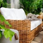 Närbild av Rossvik 2,5-sits rottingsoffa i handflätad naturrotting med tjocka vita dynor, omgiven av gröna växter och en mysig filt i ett stilfullt uterum.