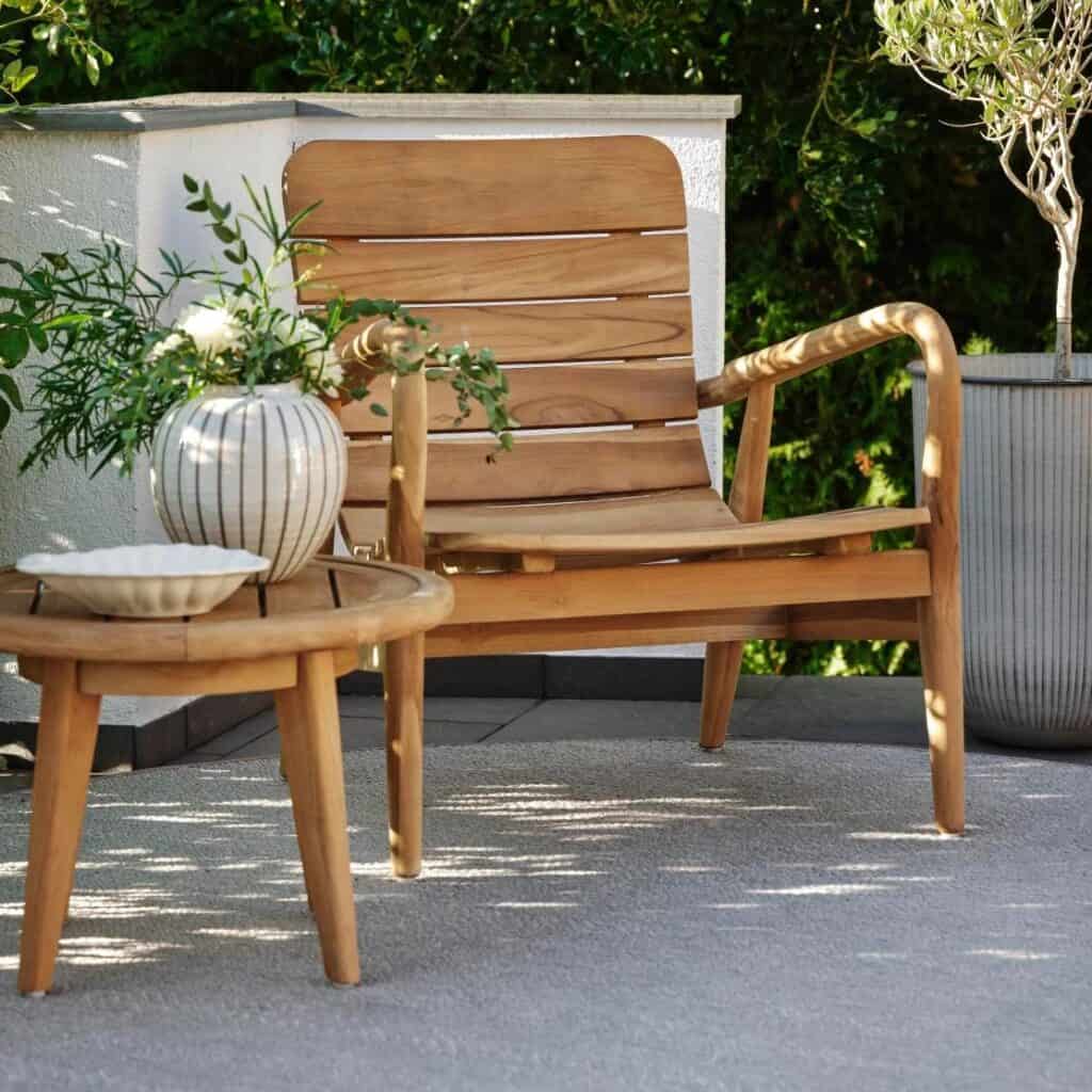 Sidobord LILJA i teak placerat bredvid en matchande loungefåtölj i teak på en grönskande uteplats, dekorerat med en keramikvas och skål.