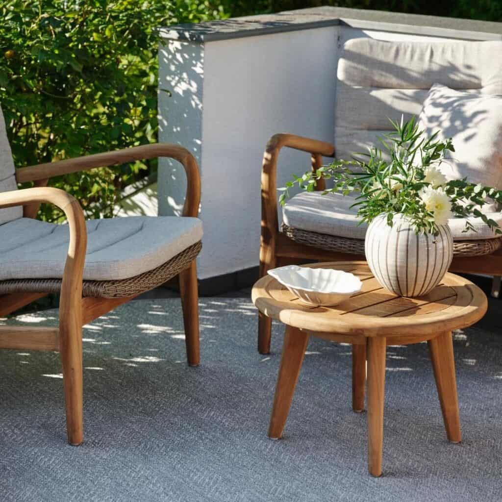 Sidobord LILJA i teak på en solig uteplats, omgiven av loungemöbler med mjuka dynor och en dekorativ vas med blommor.
